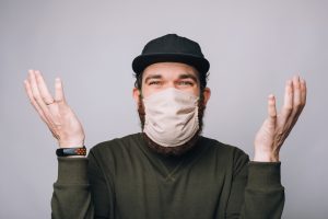 Photo of confused young bearded hipster man gesturing and wearing anti covid mask