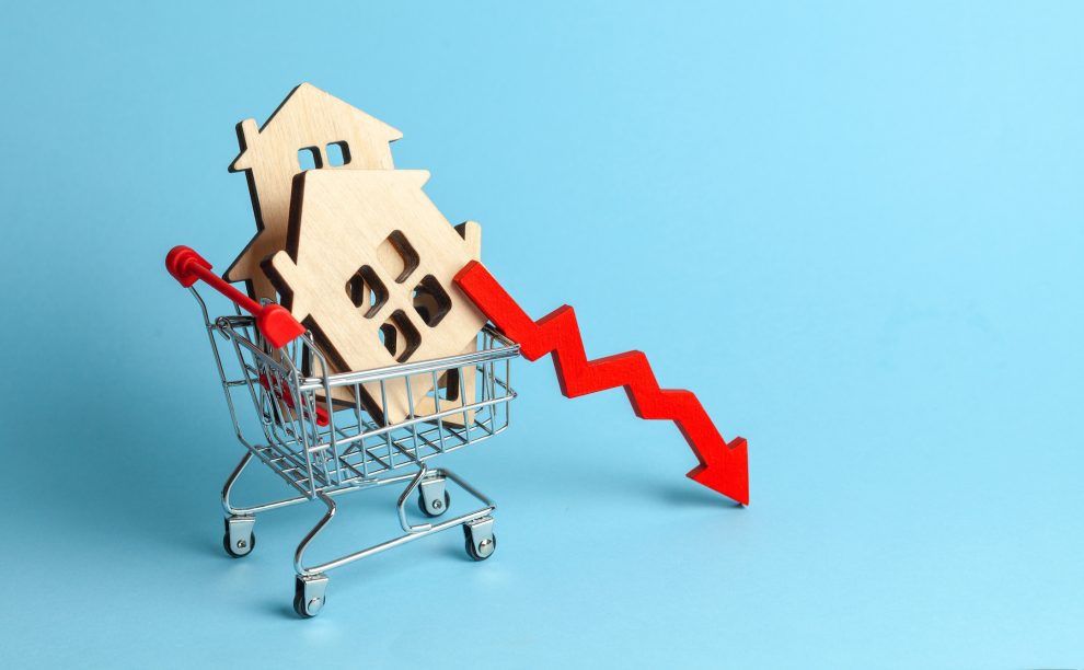 Arrow to down and houses in shopping cart on blue background. Market growth in real estate prices.